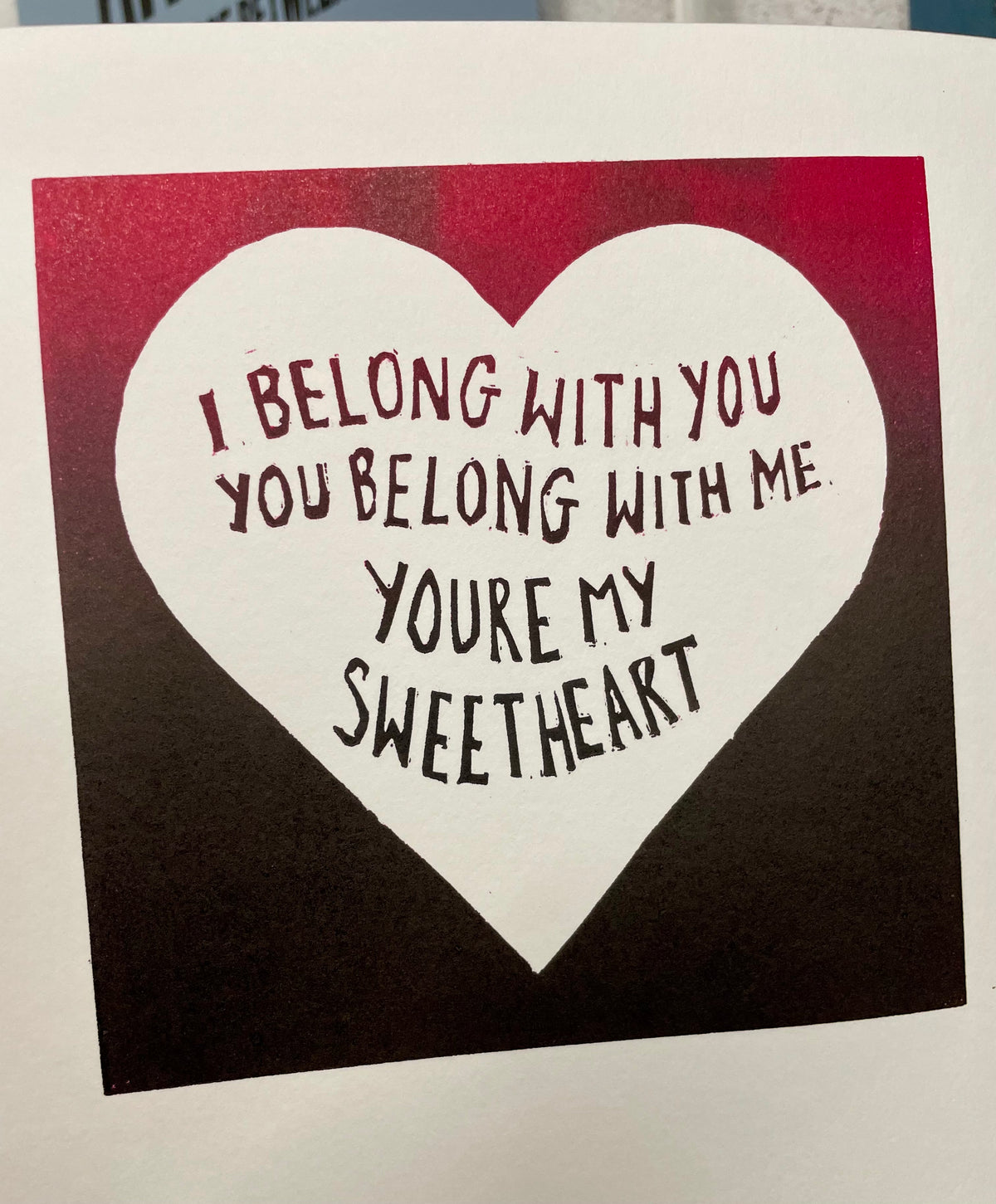 A square art print or card featuring a large white heart on a dark red-to-black gradient background, with the phrase 'I BELONG WITH YOU YOU BELONG WITH ME YOU'RE MY SWEETHEART' written inside the heart.