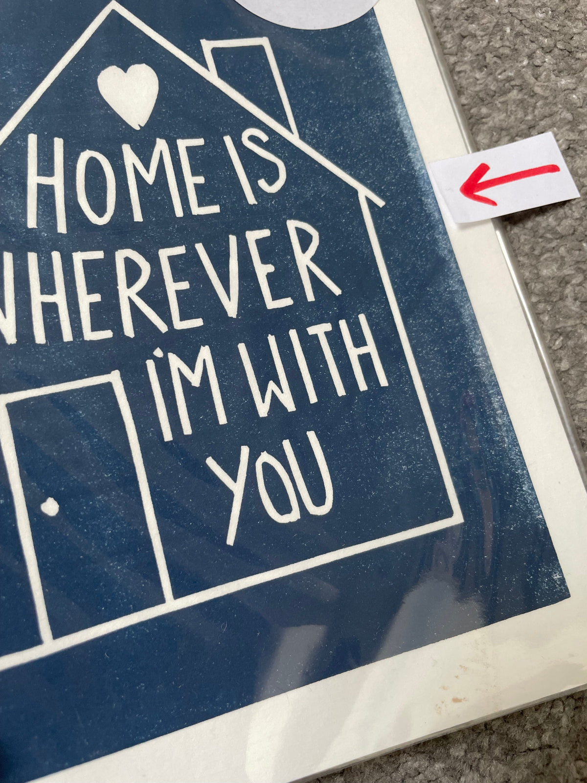 A close-up of a square wall art print featuring a navy blue house outline with white text reading 'HOME IS WHEREVER I'M WITH YOU' and a small white heart. A small white paper with a red arrow is taped near the top edge, and the print is photographed on a textured gray surface.